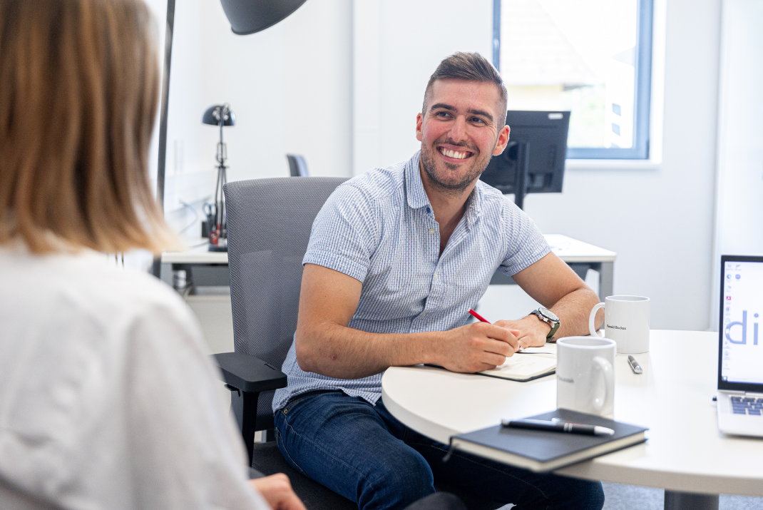 Berti sitting in the office in front of a desk while smiling at a colleague