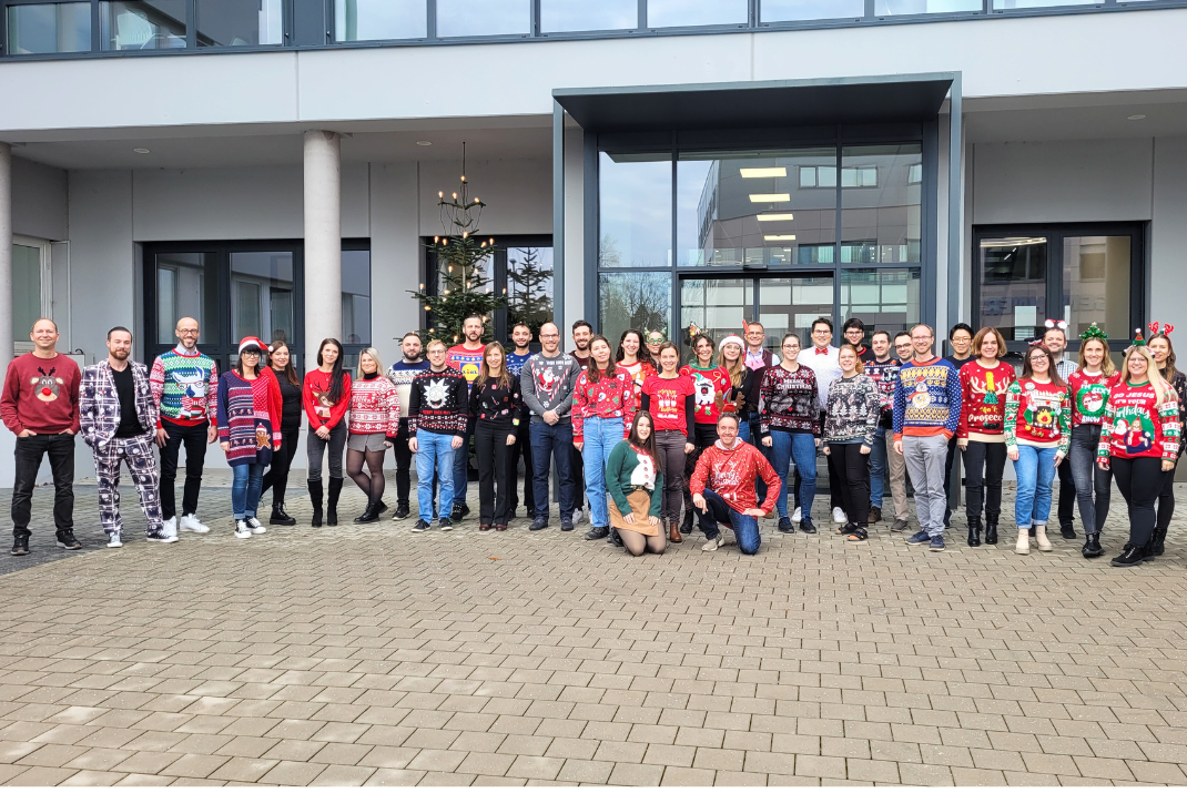 Group picture of STRATEC employees wearing Christmas sweaters in front of the entrance of the German headquarters in Birkenfeld