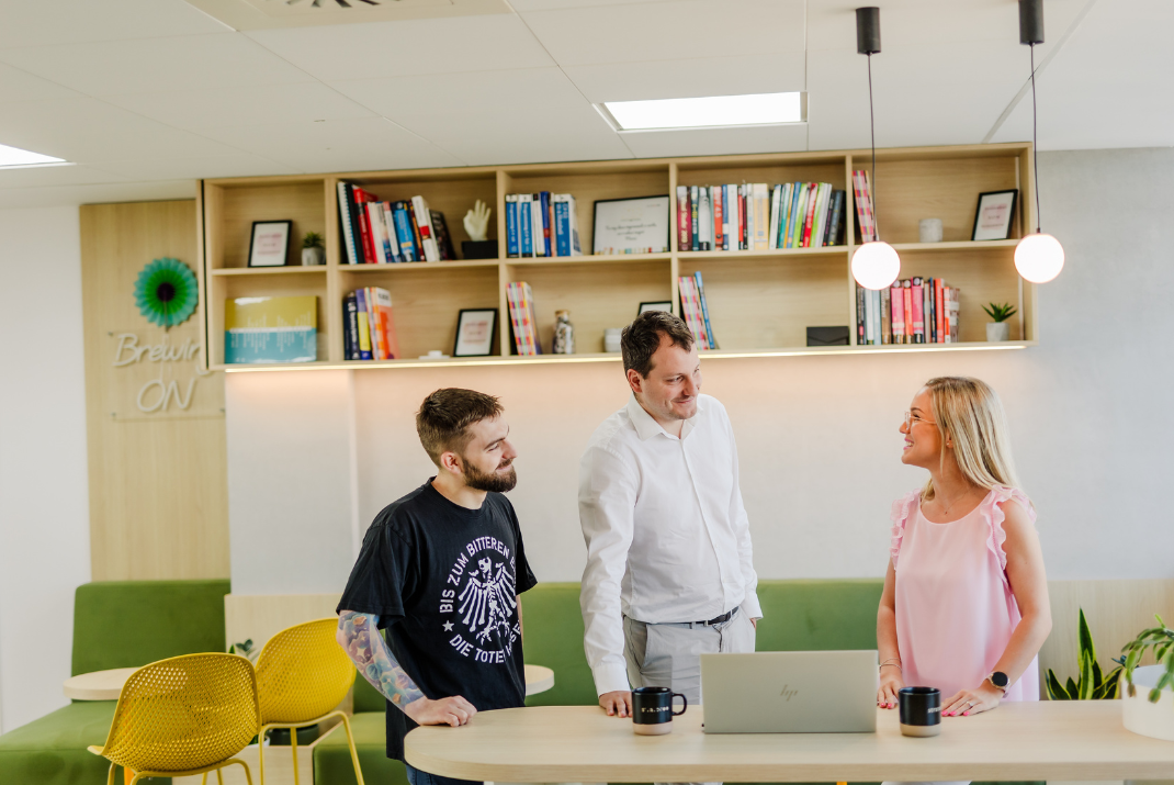 STRATEC employees in an open space office talking with each other with a small book shelf in the background